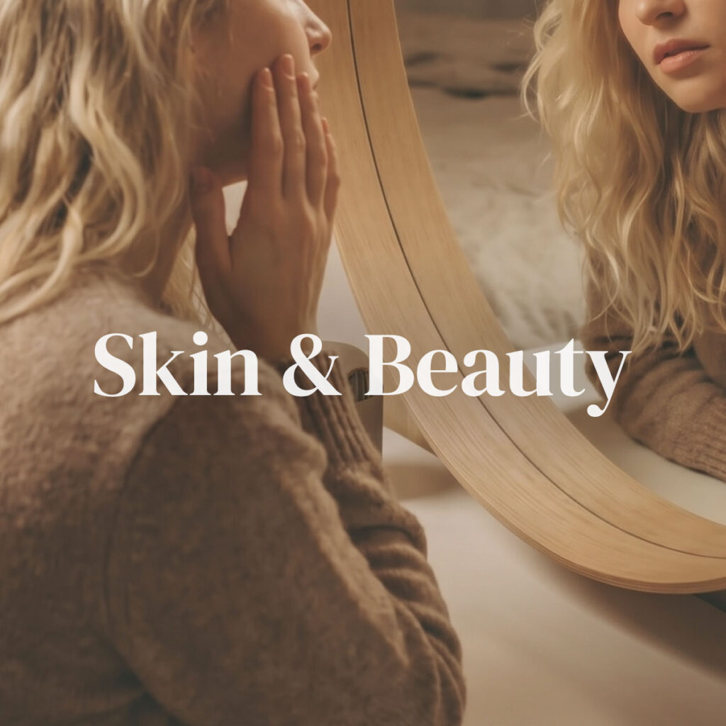 A woman gently touching her cheek while looking into a round mirror in soft natural light — representing the Skin & Beauty category focused on healthy skin, glow, and inner-out beauty care